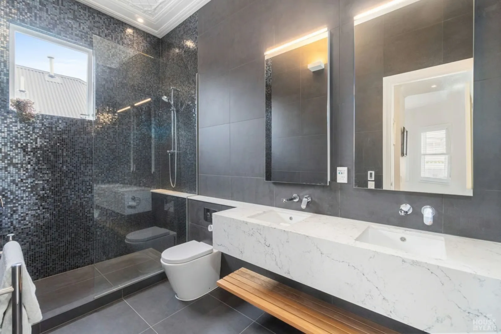 A dark mosaic tiled wall behind a stone trough basin, with a backlit mirror above it