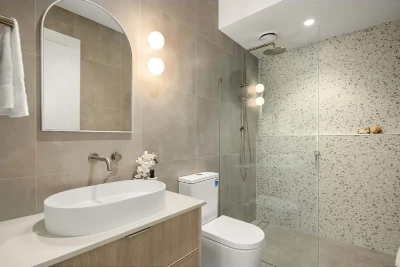A terrazzo tiled shower next to a timber vanity with an oval basin and an arched mirror